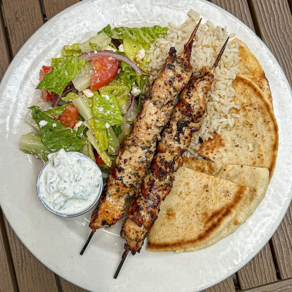 Grilled chicken skewers with rice, a side salad, pita bread, and tzatziki sauce on a white plate.