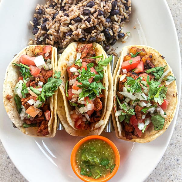 Three tacos with meat, diced tomatoes, onions, cilantro, and a side of rice, beans, and salsa.