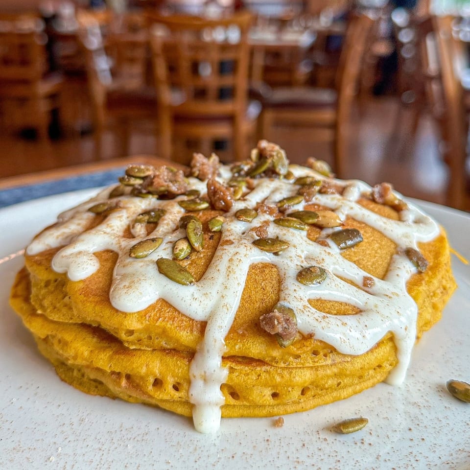 Stack of golden pancakes with white icing, pumpkin seeds, and cinnamon on a white plate.