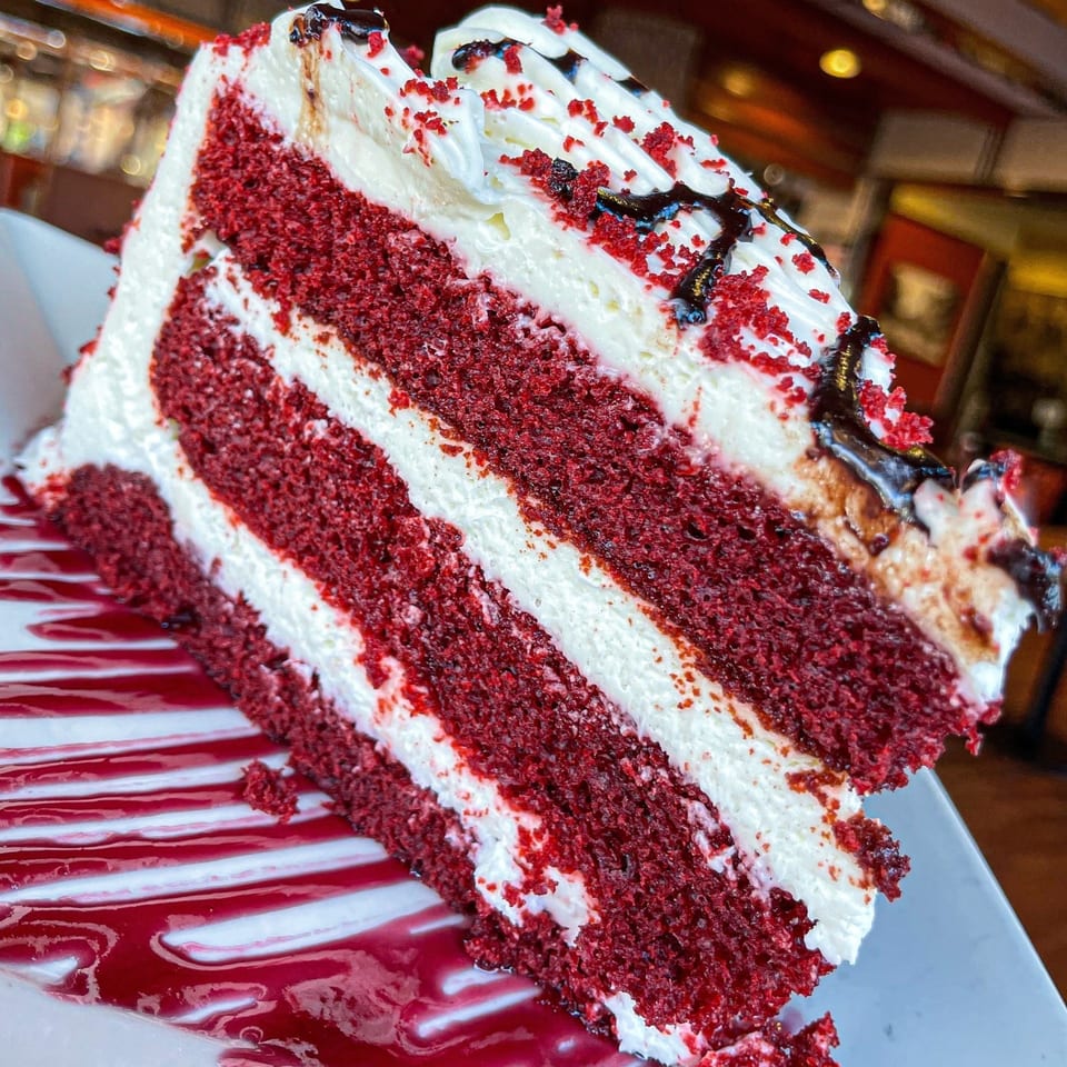 A large slice of red velvet cake with white frosting, chocolate drizzle, and red sprinkles.